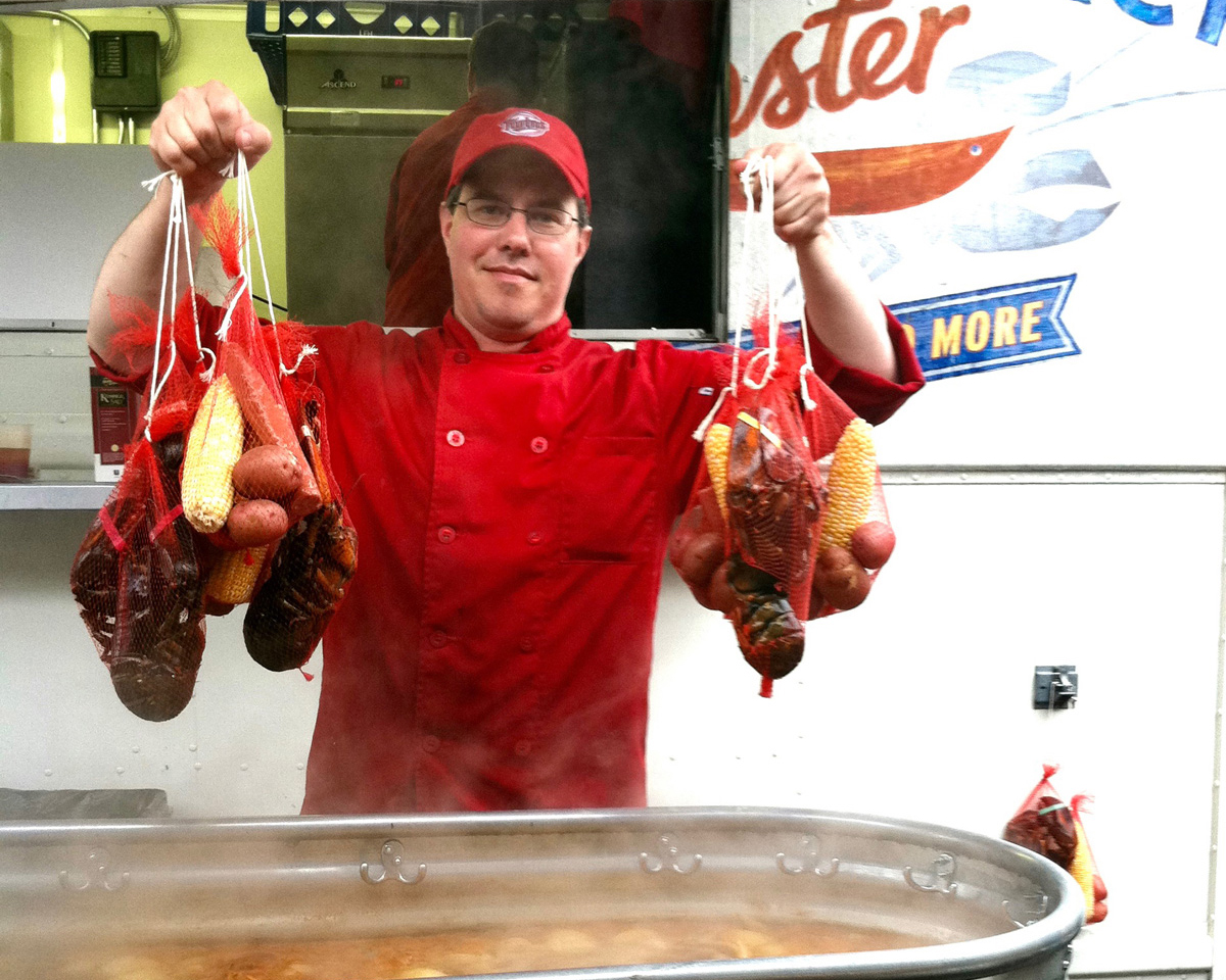 Smack Shack owner Josh Thoma in a red chef coat standing in front of the food truck, holding bags full of seafood for a boil